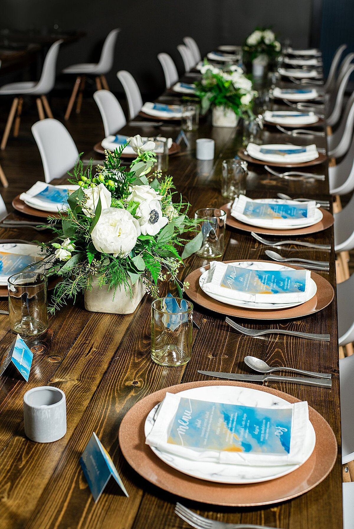 Long brown farm table set with silver flatware, copper chargers,  marbled plates, a white folded napkin accented with a teal and gold menu card. A long white and green centerpieces run down the center of the table.