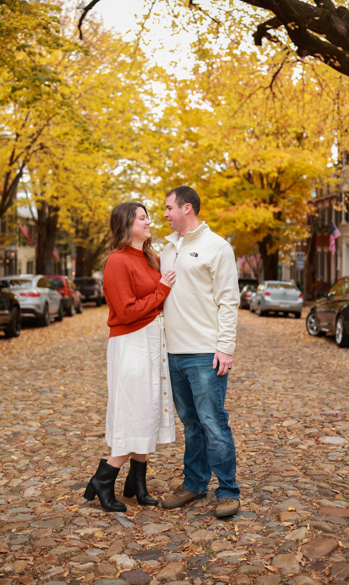 man and woman standing next to each other with yellow trees in the background and standing on pebble stones