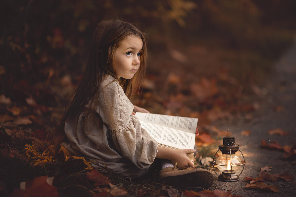 A little girl sits on the groundwith a book in her lap and glowing lantern beside her in Kamloops