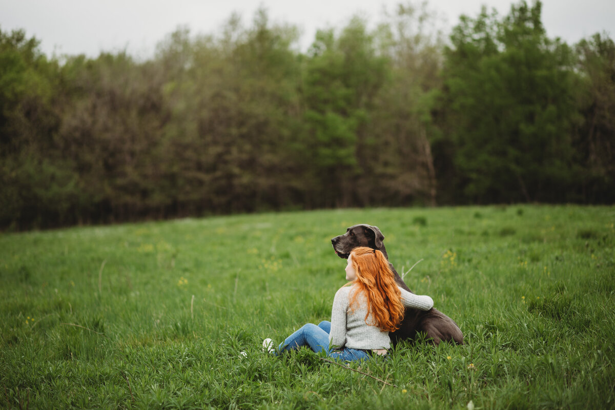 A red headed girl leans up against a gray dog with her arm wrapped around it. They sit in the middle of a grass field looking off into the distance together.