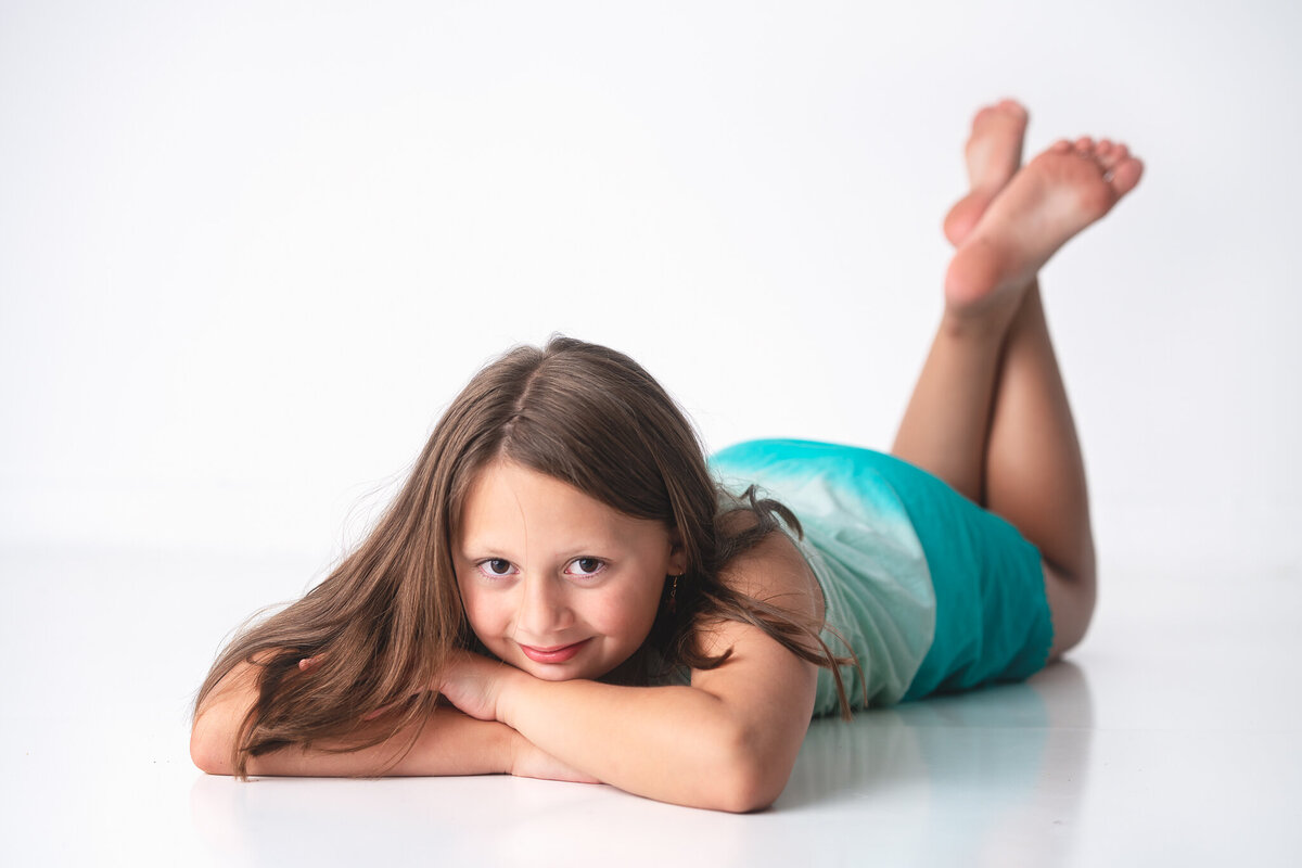 A young girl in a blue dress is lying down in a photography studio and smiling at the camera.