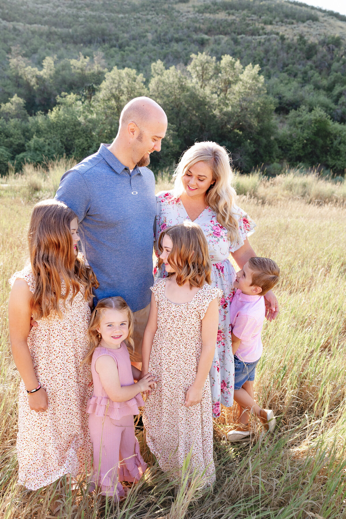 A father and mother share a loving moment with their four children in a golden field. The parents gaze at each other with smiles, while the children look up at them, capturing a heartwarming family connection
