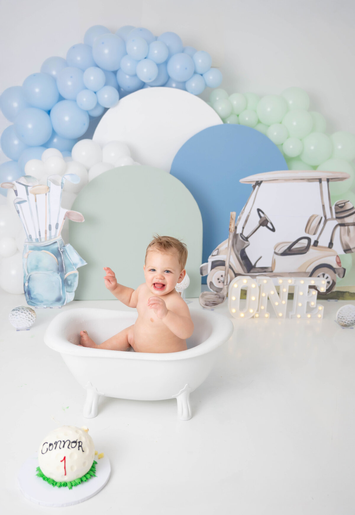Baby laughing while sitting in a mini tub surrounded by blue, green and white arches with coordinating balloons. 