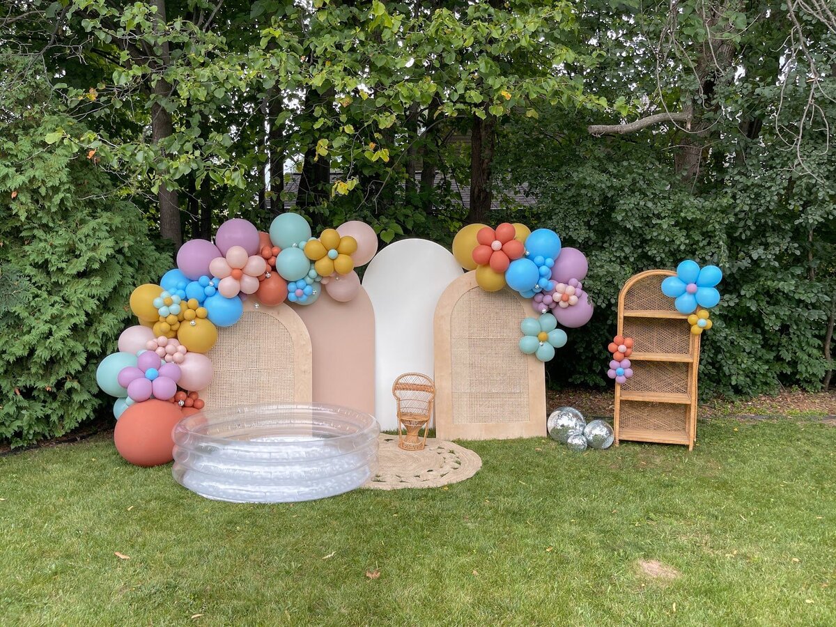 Yellow, blue, purple, and pink balloons attached to four wooden arch backdrops and a rattan shelf. A clear ball pool and toddler size peacock chair set up in front.