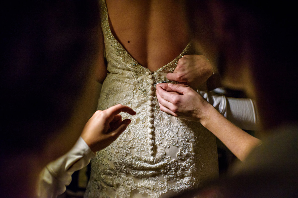 A mother and sister help a bride button up her lace wedding dress.