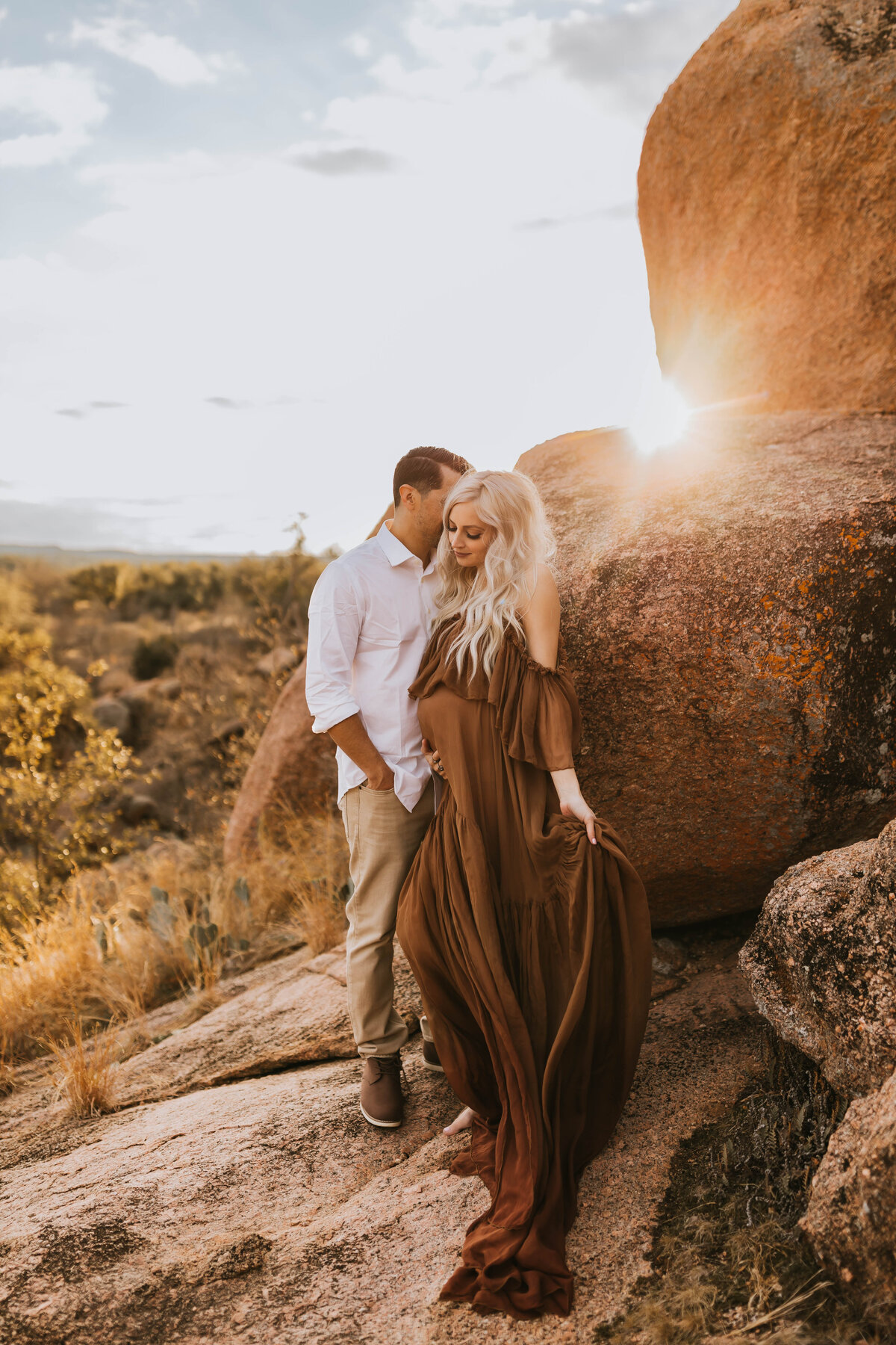 expecting couple getting close on a boulder with the sun peeking through at the Wichita Mountain Wildlife Refuge