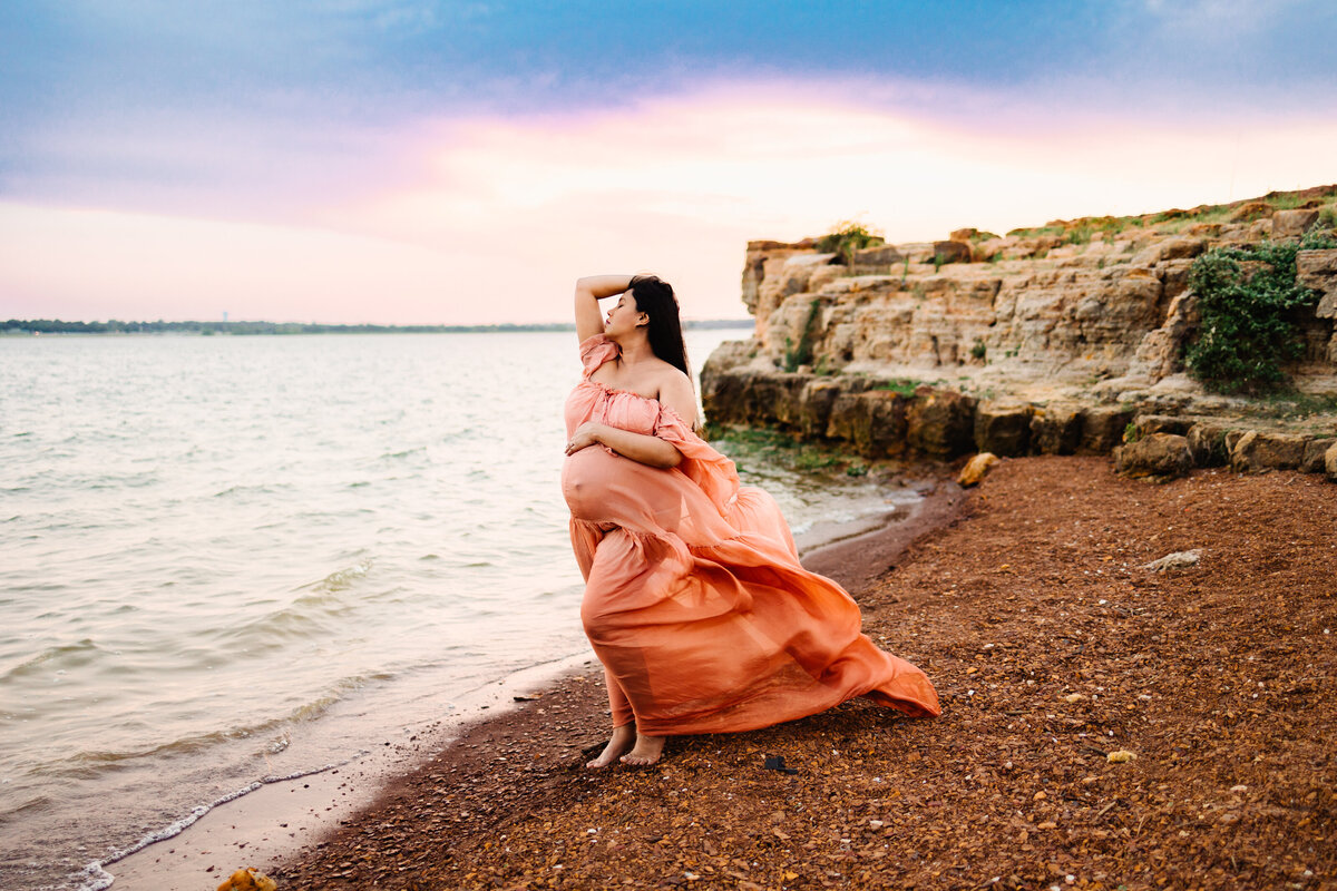 Maternity photography in Albuquerque featuring a pregnant woman in a flowing orange dress standing against a stunning beach backdrop. The scene includes the sea, sand, and mountains under a clear blue sky. The woman has her hand on her head and her eyes closed, capturing a serene and graceful moment.