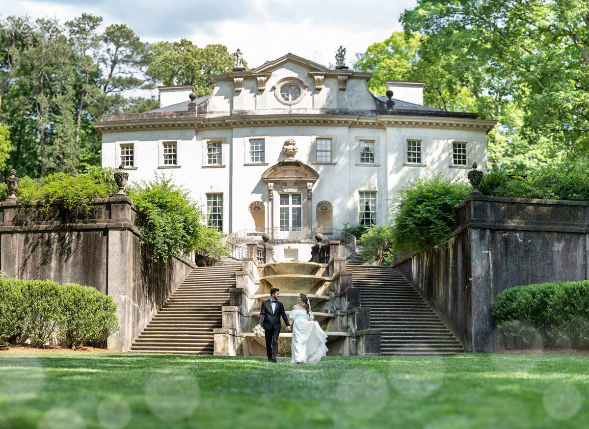 This photo shows a happy couple walking down the stairs of a majestic white estate, surrounded by lush greenery and sunlight. Photographed by GreenPoint Photograph.