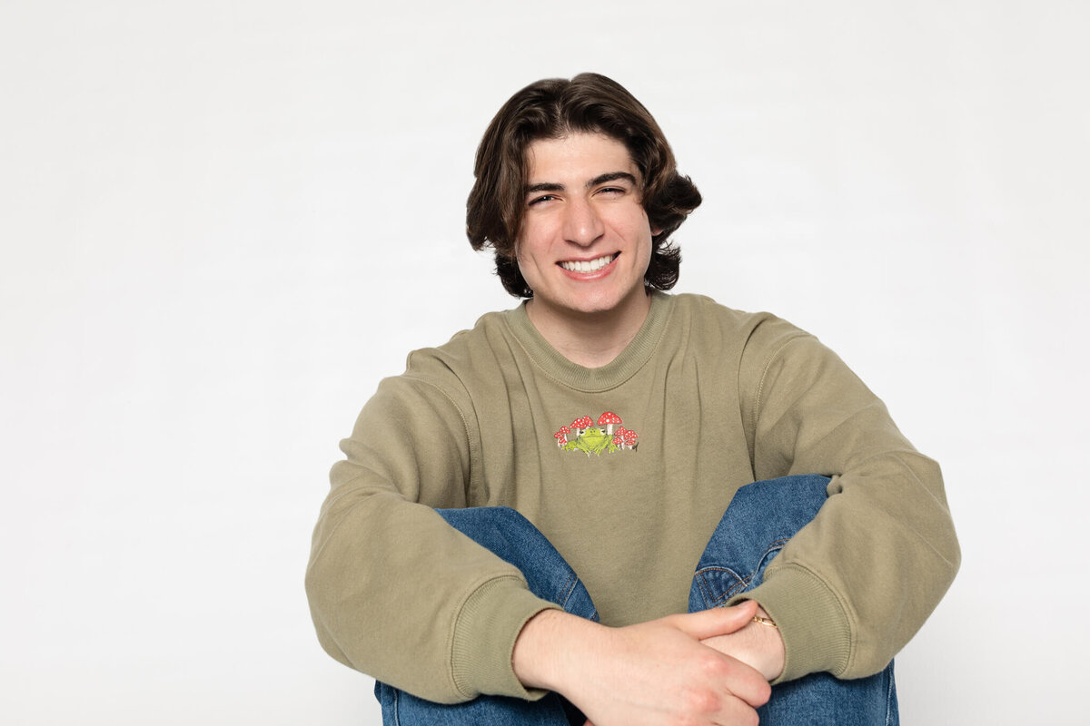 A person with long dark hair and a warm smile sits casually against a plain white background, wearing a green sweater with a colorful logo and blue jeans. Their posture is relaxed, with arms resting on their knees.