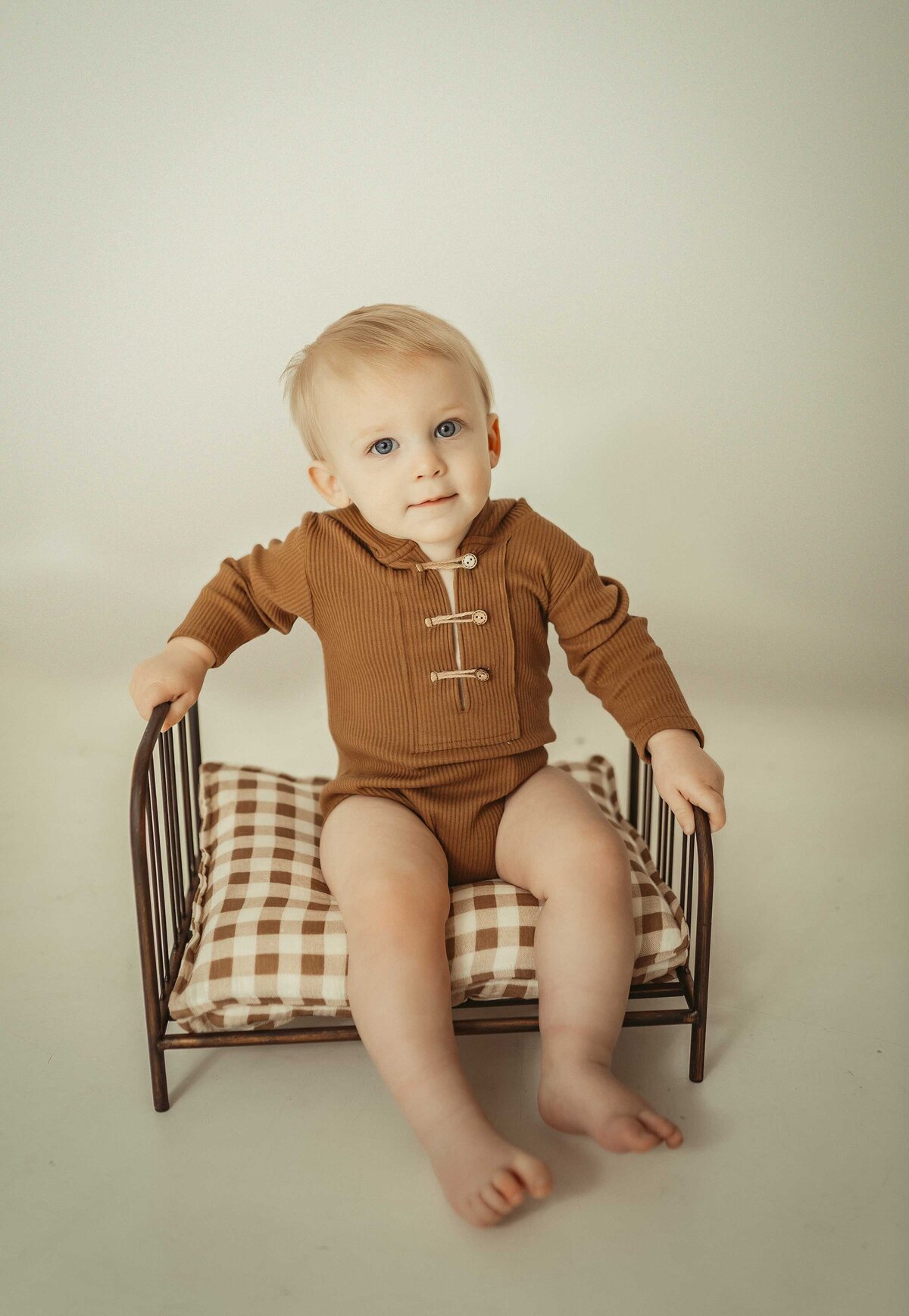 young boy sitting on a toy metal crib wearing a brown outfit