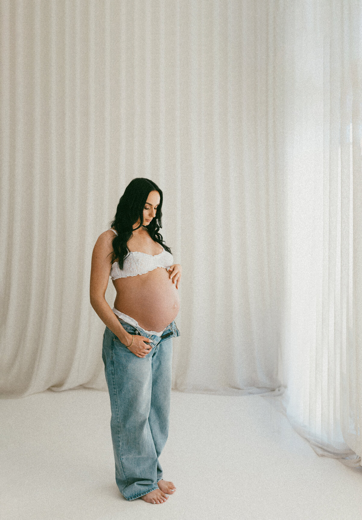 A pregnant woman standing in a white top and jeans and looking down at her belly during a studio maternity photoshoot