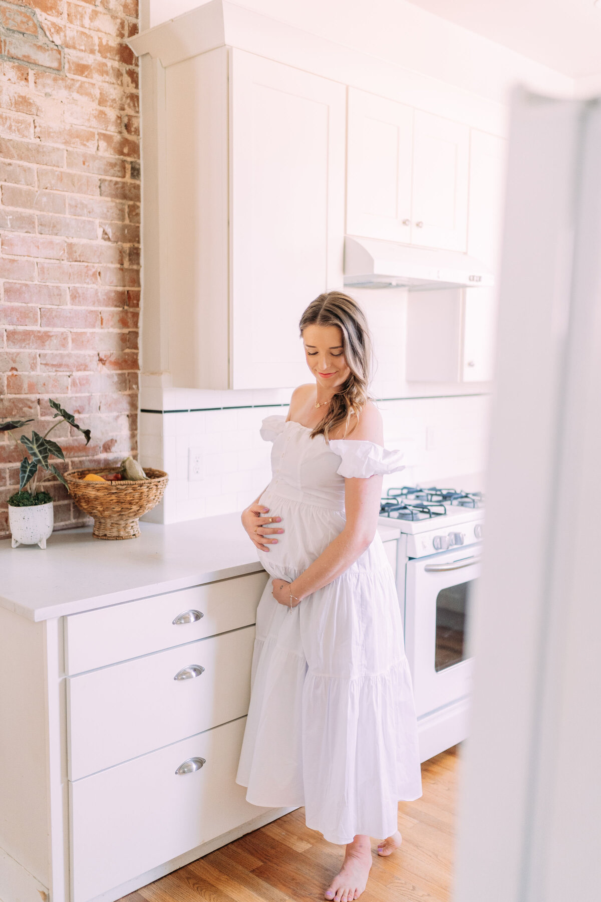mother in white dress tenderly holds belly while standing in kitchen oklahoma