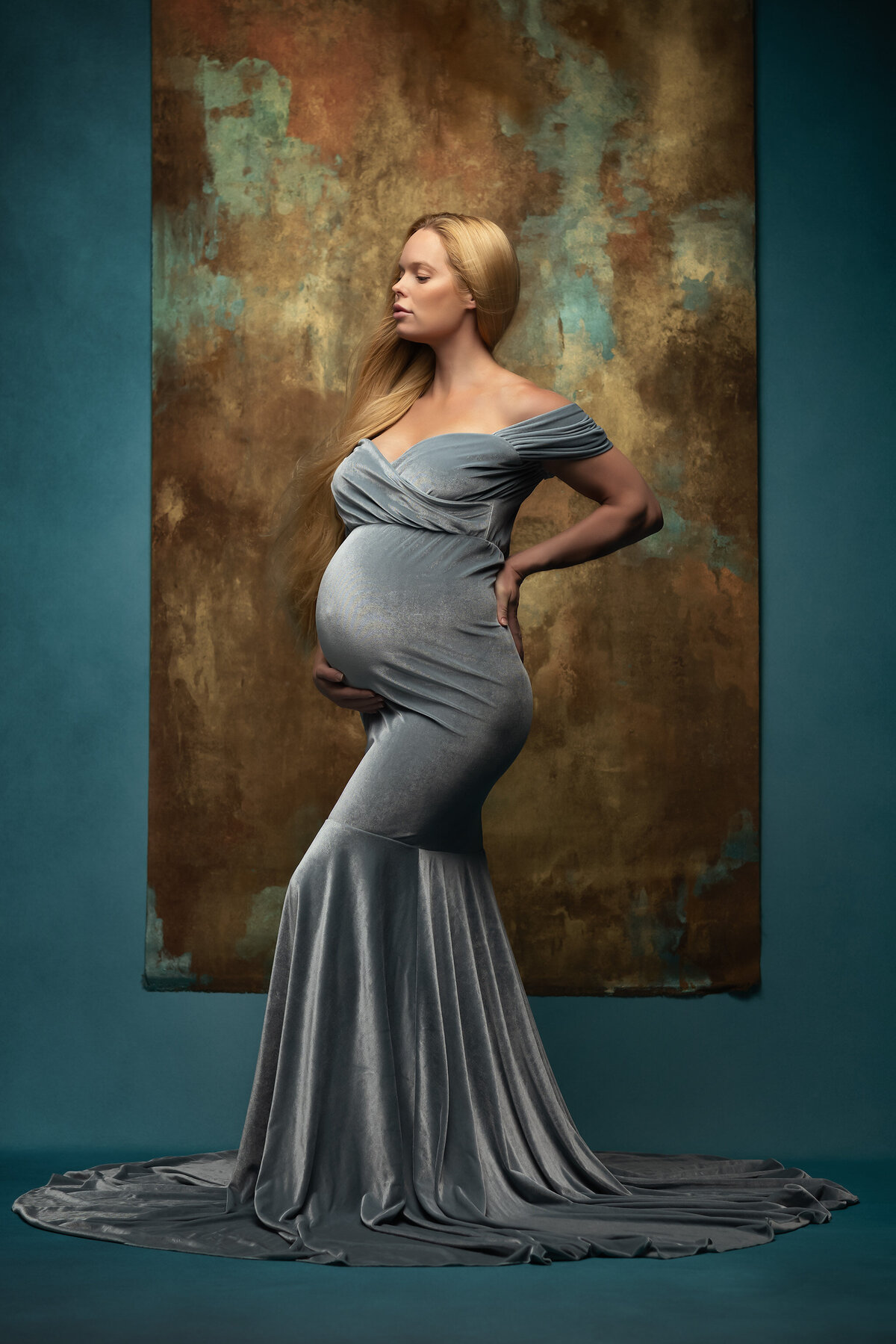 A stunning portrait of an expecting mother in a flowing, blue velvet gown, standing gracefully against a rich, textured gold and teal backdrop. Her pose is elegant, with one hand gently supporting her belly, and her long blonde hair cascades down her shoulder.