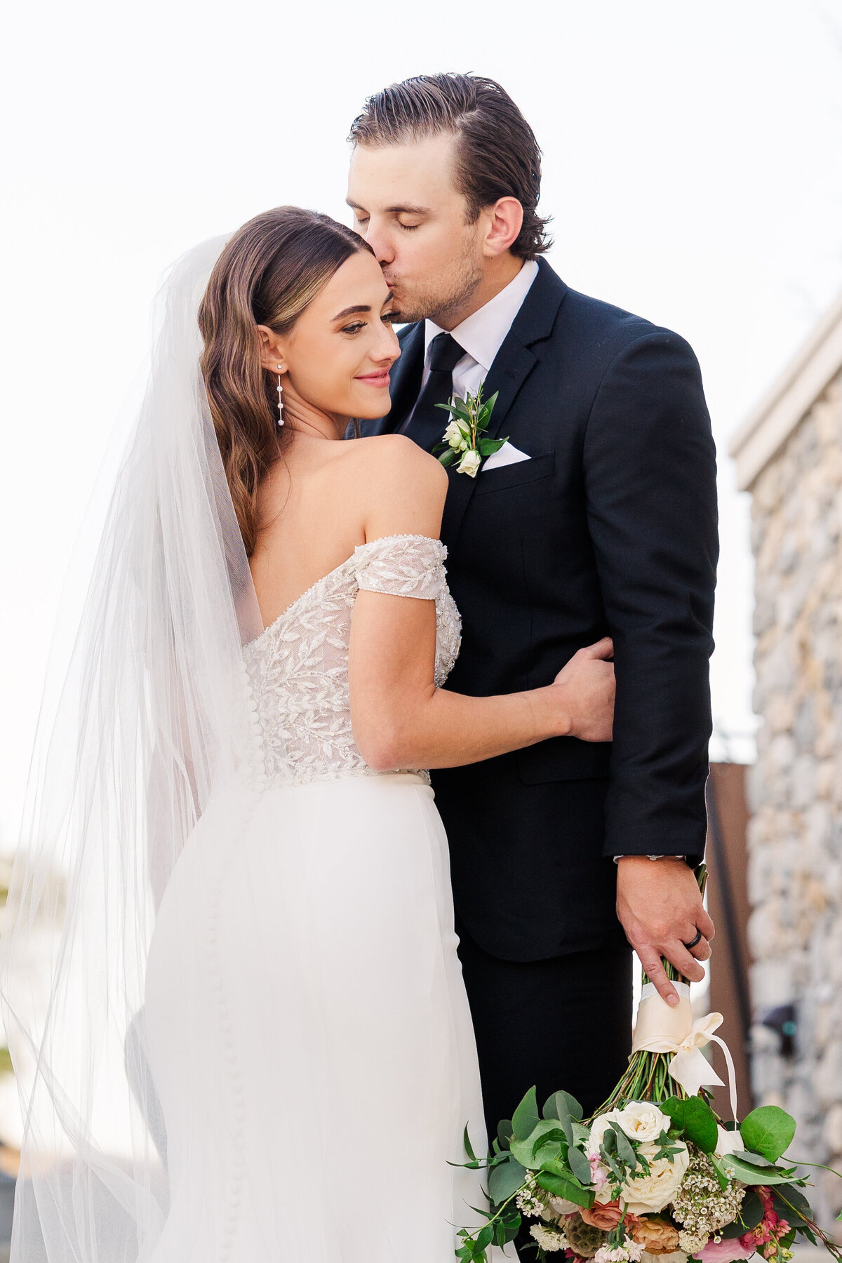 A graceful moment between bride and groom as they embrace near stone architecture, the bride’s bouquet softly resting in hand. This refined portrait was taken in Westlake, Texas and embodies upscale romance for DFW, Dallas, and Fort Worth wedding celebrations.
