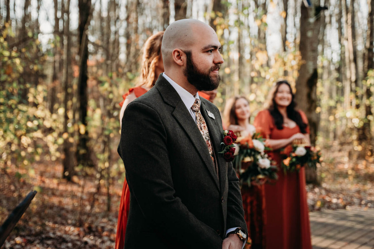 Groom watches their partner walk toward them in fall trees