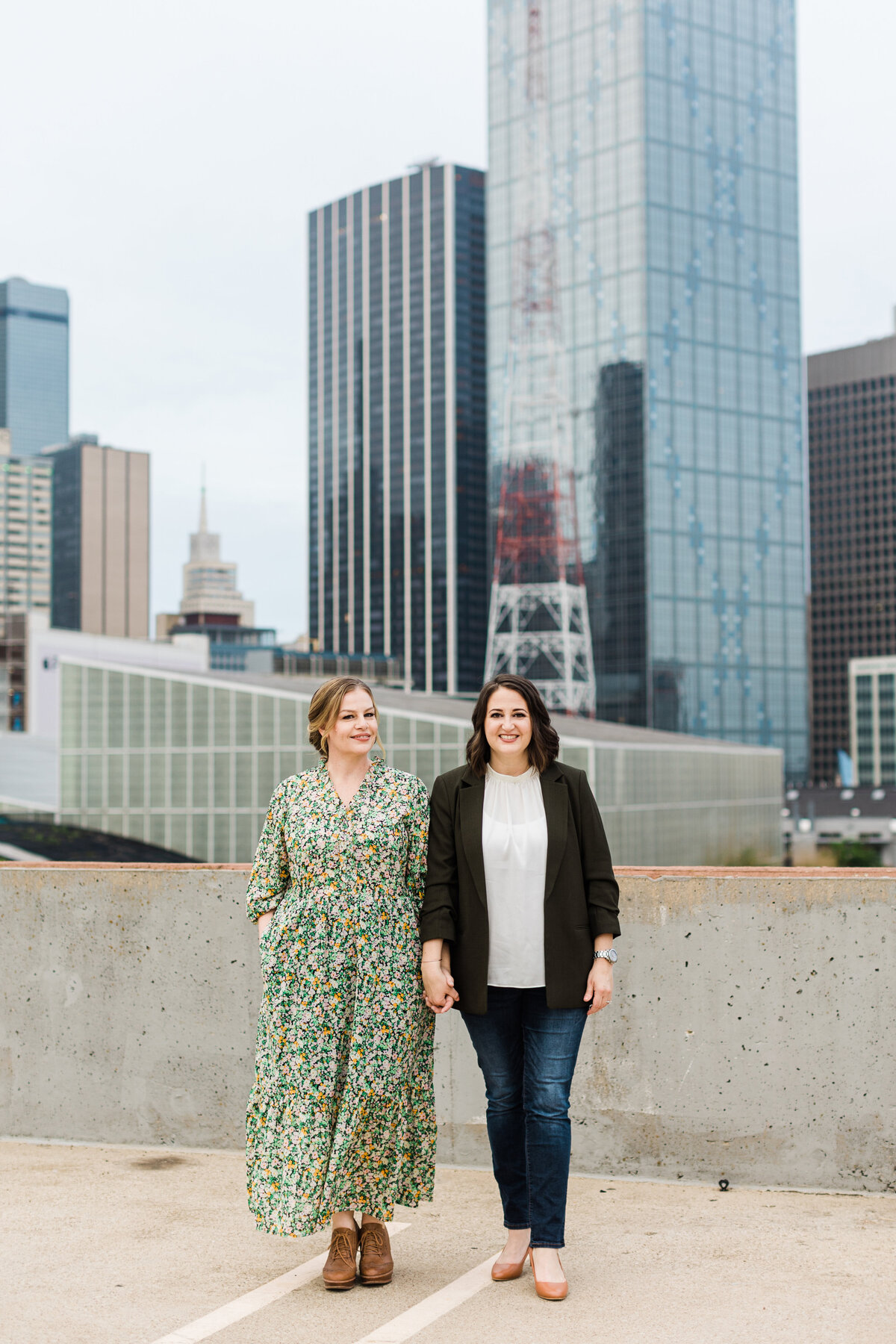 Two women holding hands and posing on top of a parking garage during their downtown engagement photoshoot in Dallas, Texas.The woman on the left is wearing a long, colorful floral dress and brown shoes. The woman on the right is wearing a white shirt, black blazer, jeans, and heels. A portion of the Dallas skyline can be seen towering behind them.