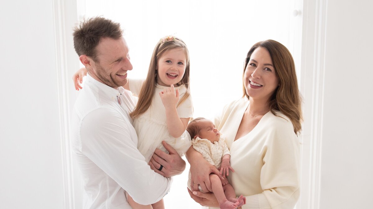 A family of four stands in a bright, naturally lit room at Dandelion Studio in Moira, Northern Ireland. The father holds a smiling young daughter in a light-coloured dress, while the mother cradles a newborn dressed in lace. The family is dressed in coordinating soft tones, and the scene captures a joyful, intimate moment full of warmth and connection.