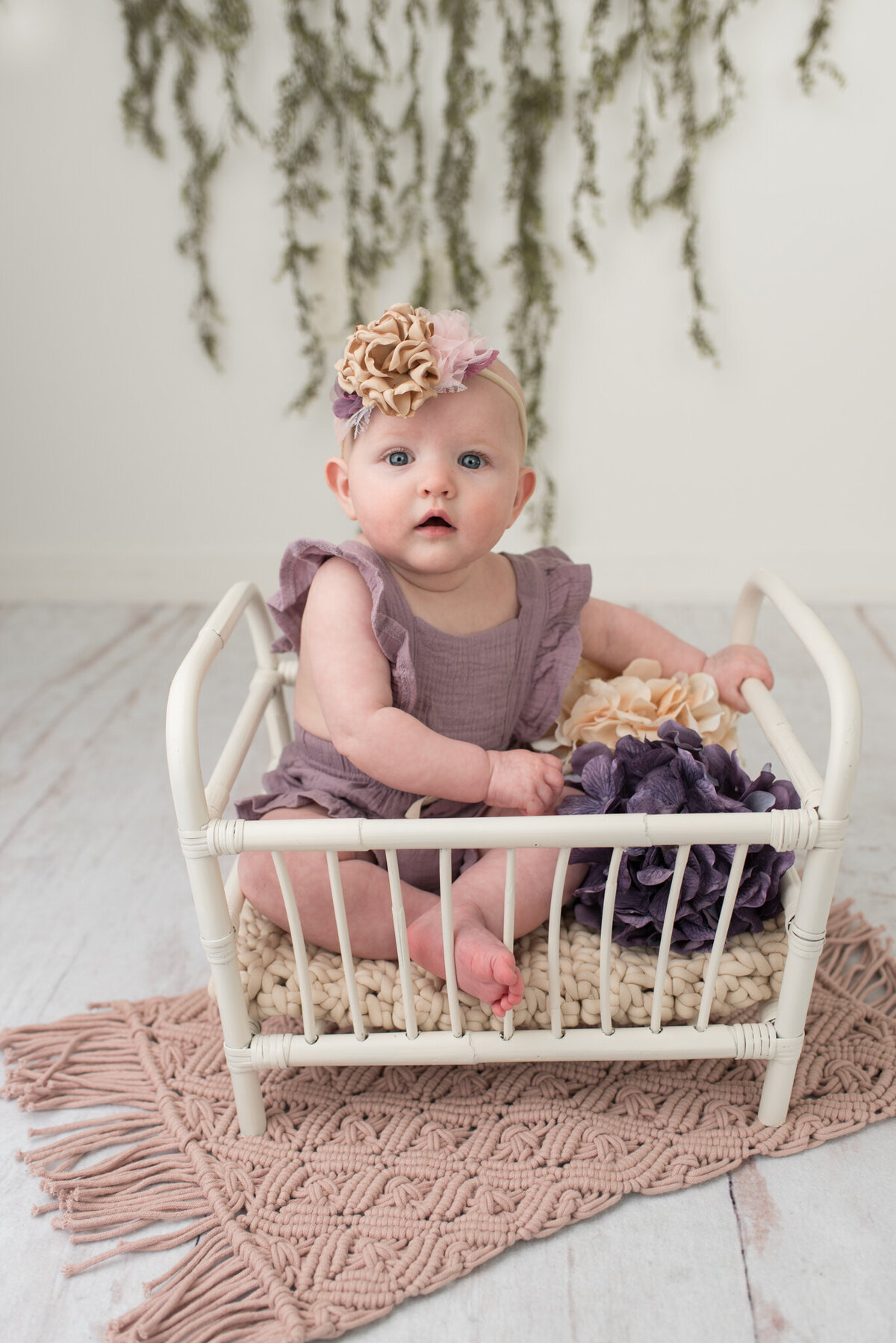 Infant girl sitting in a white crib with a purple romper and a floral headband