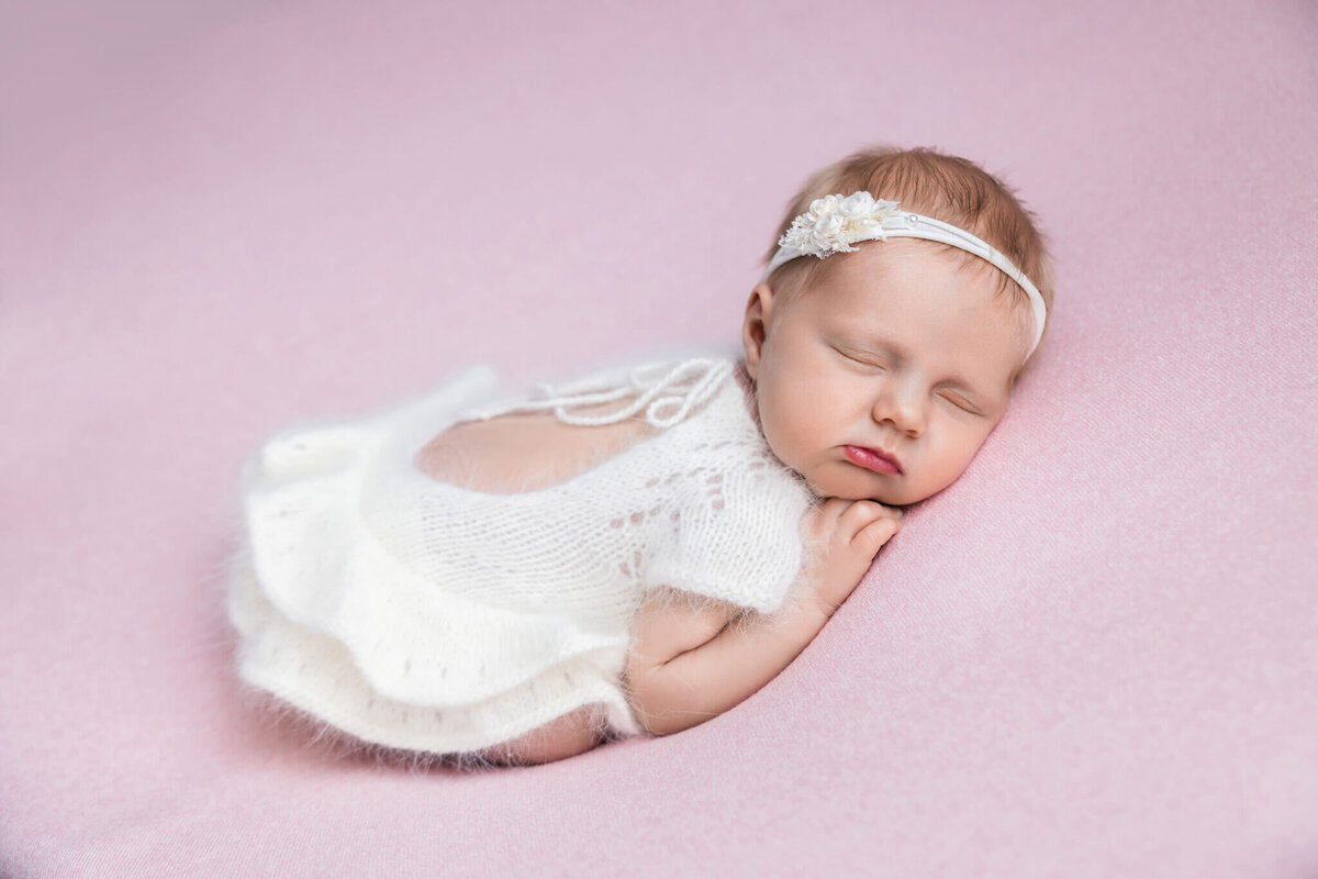newborn baby asleep in a white romper laying on a pink fabric with a white headband