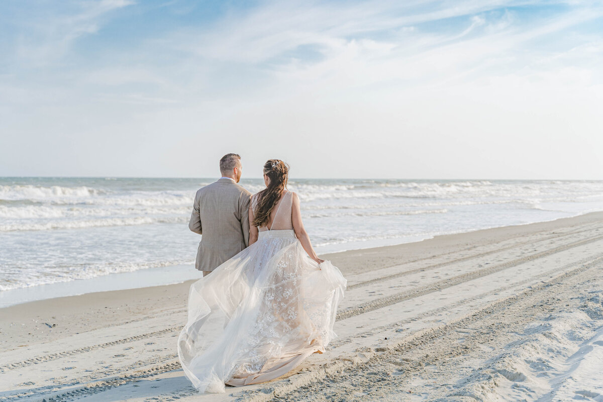 A bride and groom walk away from the camera down the beach at sunset