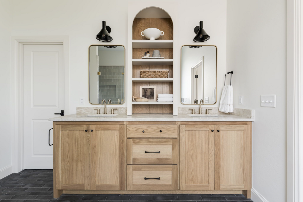 Double vanity with natural white oak cabinetry, dual brass faucets, arched open shelving, and matte black sconces above brass-framed mirrors on dark brick tiled flooring.