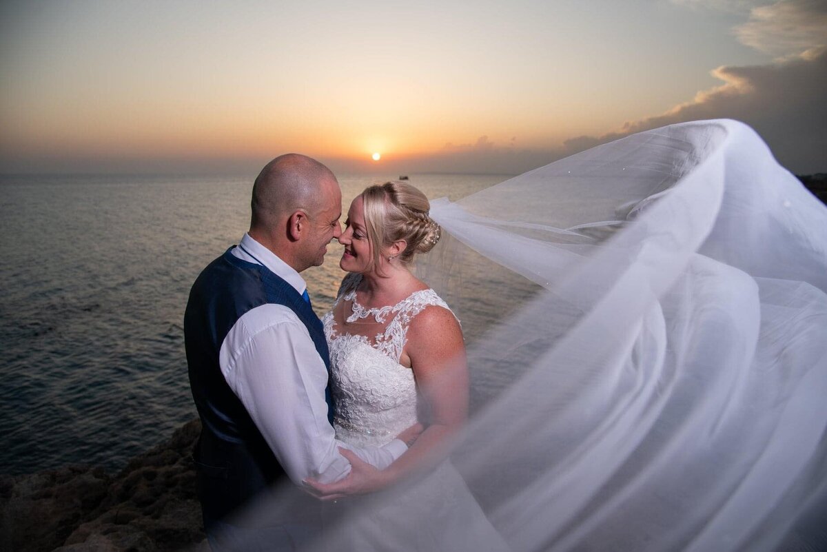 Bride & Groom stand in front of a sunset while the brides veil blows beautifully in the wind