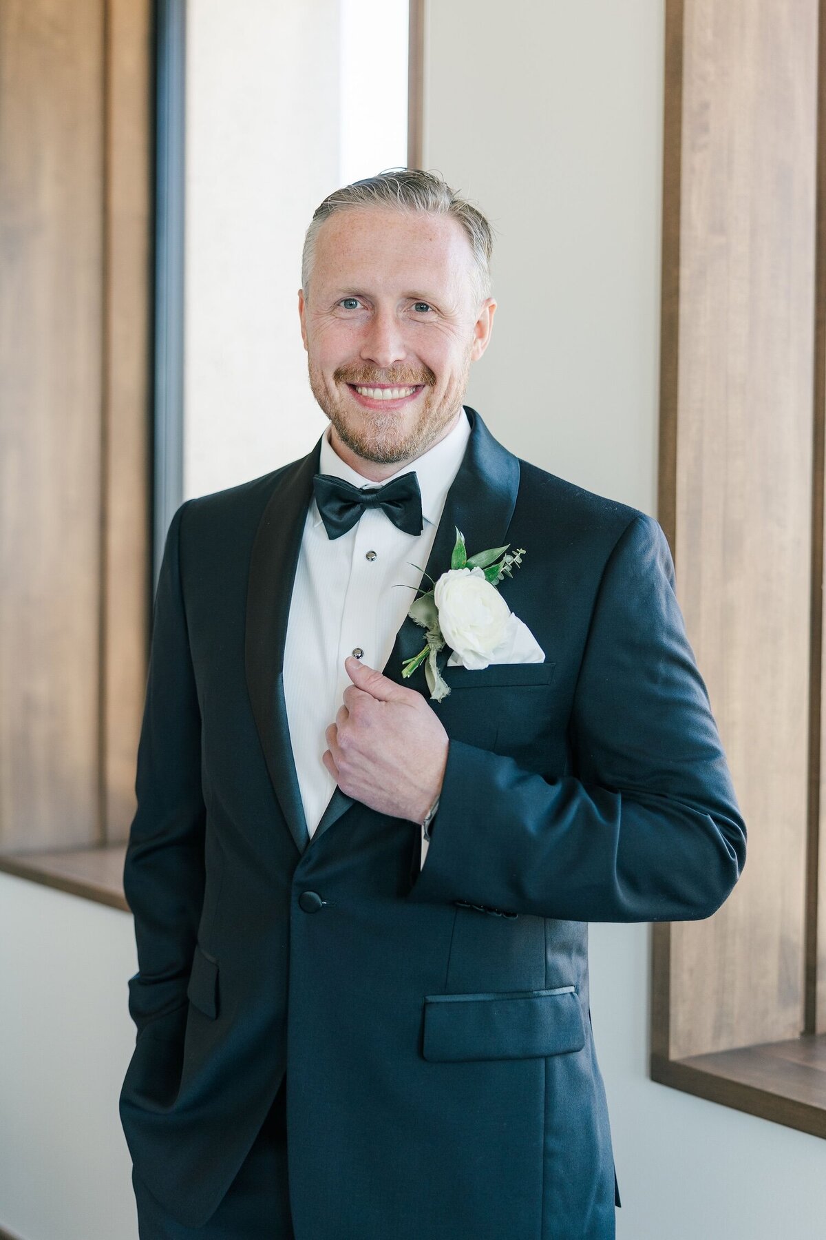 A groom stands near windows at Hotel Marcel holding his hand on his lapel. 