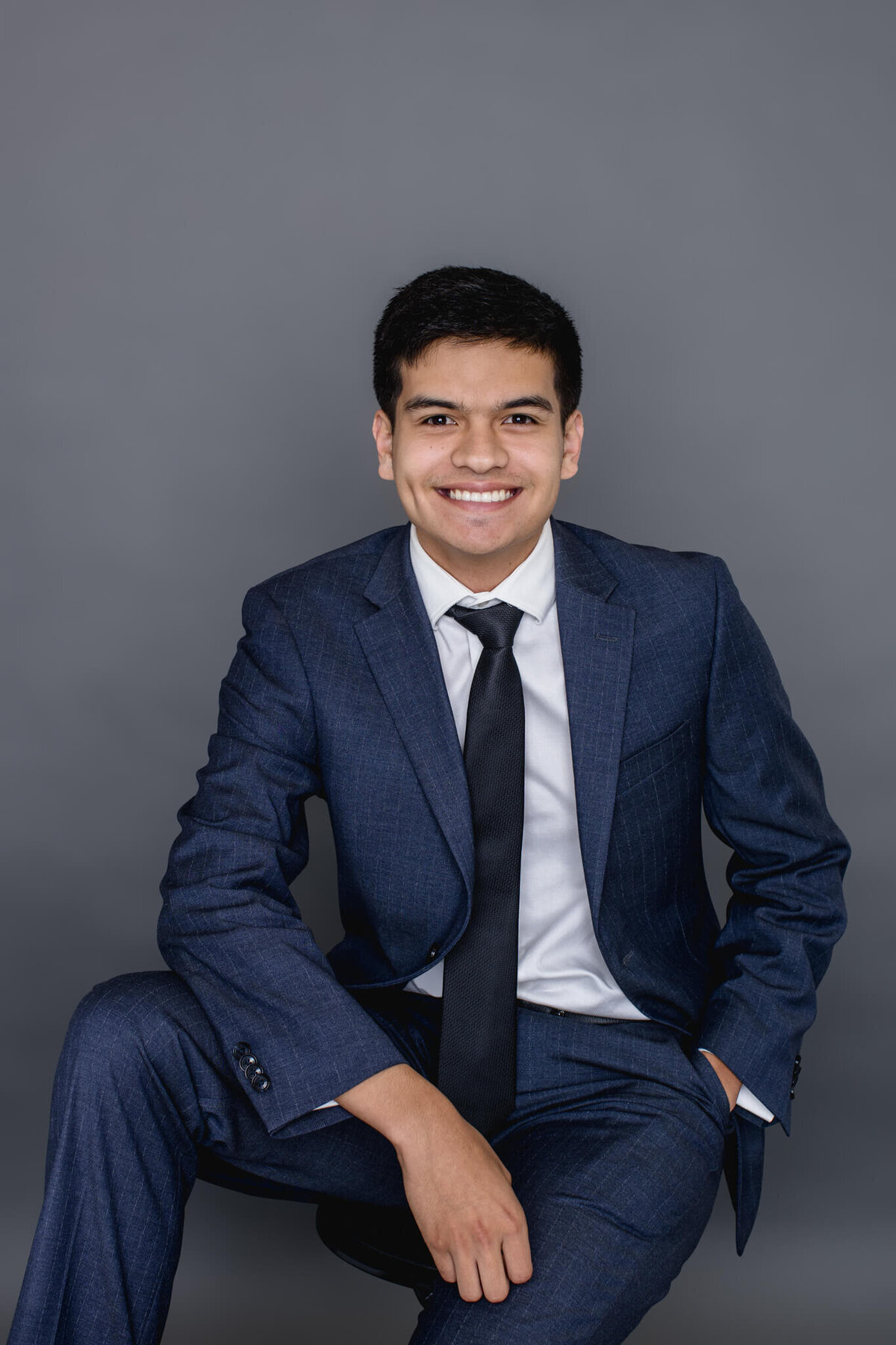 A young man with black hair wearing a navy suit jacket and dress pants sitting with his left hand in his pocket and the other arm draped over his right leg which is propped up, smiling at the camera in front of a dark grey background in a studio in Tustin.