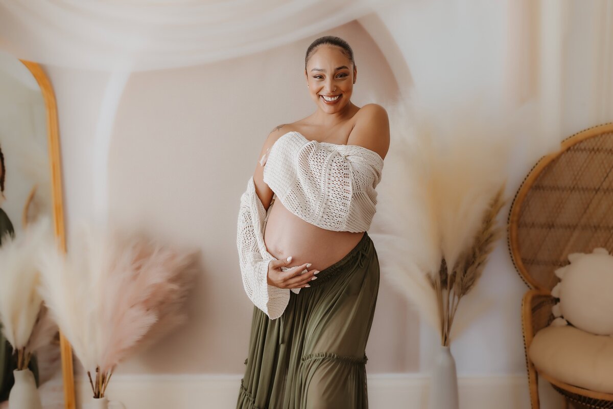 Pregnant woman smiling, holding her belly, wearing a knitted shawl and a long skirt, standing in a softly lit room with decorative pampas grass and a wicker chair.