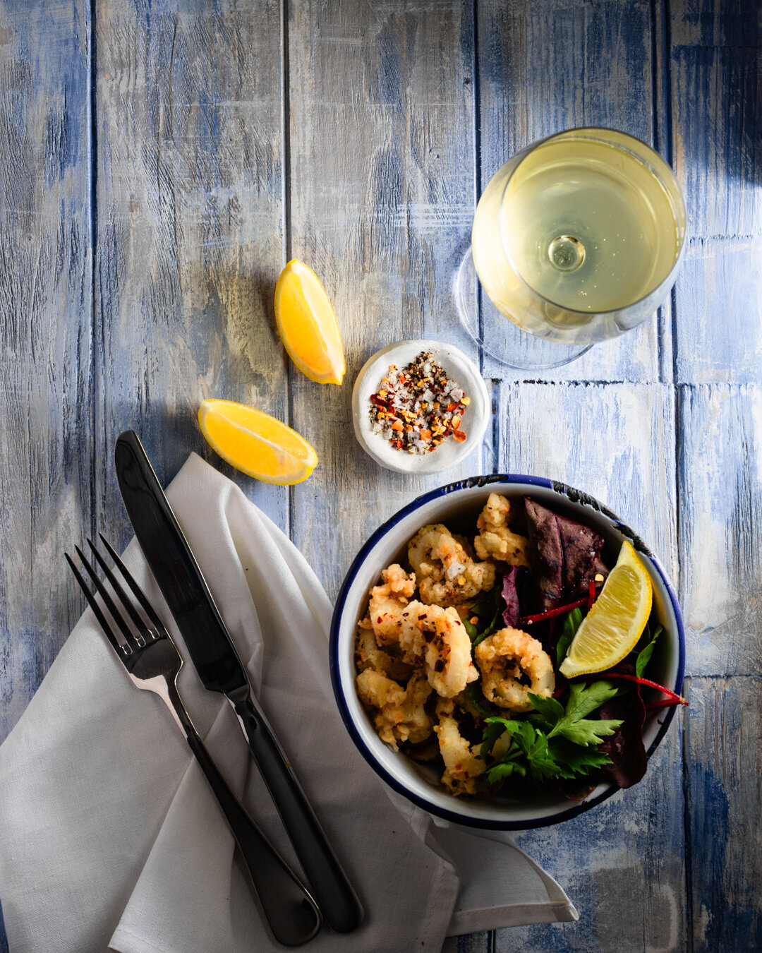 A bowl of deep fried Calamari with salt and chilli seasoning on a restaurant table with napkin, cutlery and glass of white wine with lemon wedges