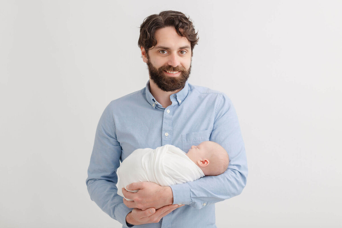 Dad in white button-down smiling at the camera while holding his baby in his arms. Baby is swaddled in a white blanket.