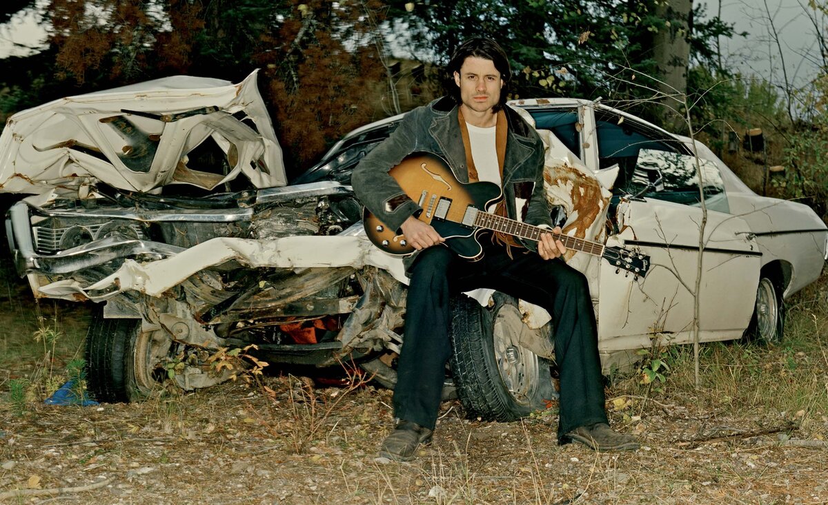 Male musician portrait Dustin Bentall sitting against smashed white car holding guitar