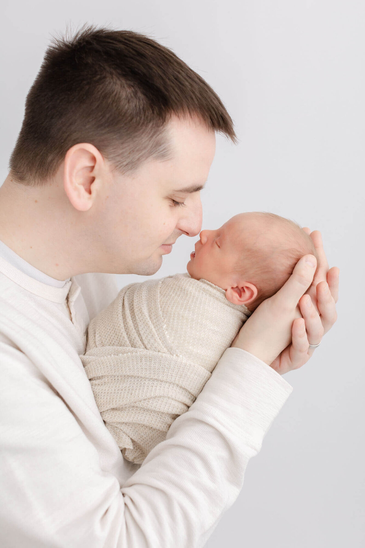Daddy holding baby in his arms and touching noses together. Baby is sleeping peacefully. Dad is wearing a light cream shirt and baby is wrapped in a beige wrap. Image taken during newborn portrait studio in minimalist newborn photography studio. Photo taken by Ashlie Behm Photography