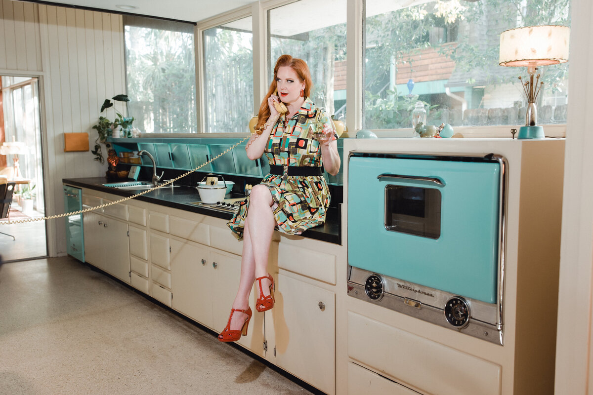Pinup model poses in a colorful midcentury kitchen, wearing a geometric dress and heels while talking on a vintage rotary phone.
