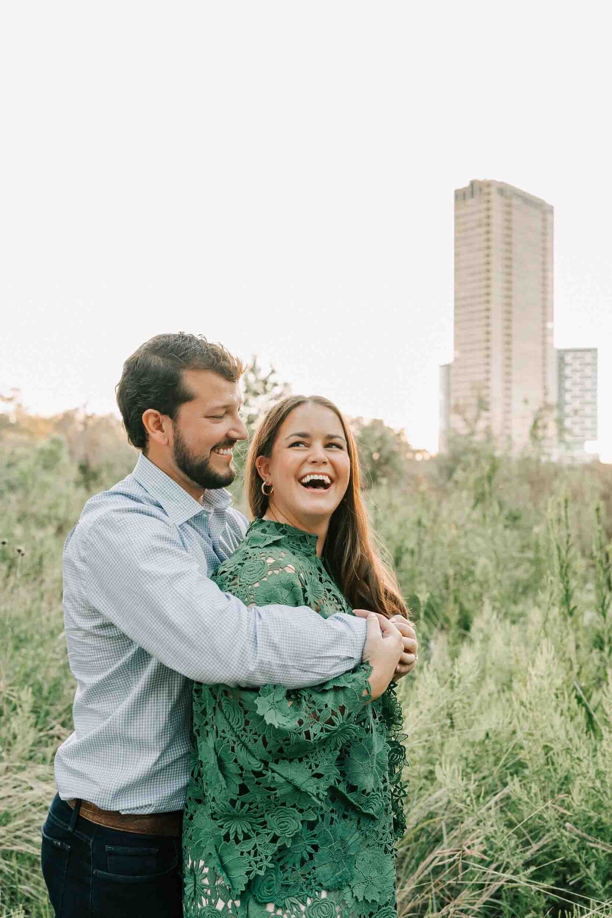 kimberly and garrett take engagement photos with houston skyline behind them while he holds his arms around her.