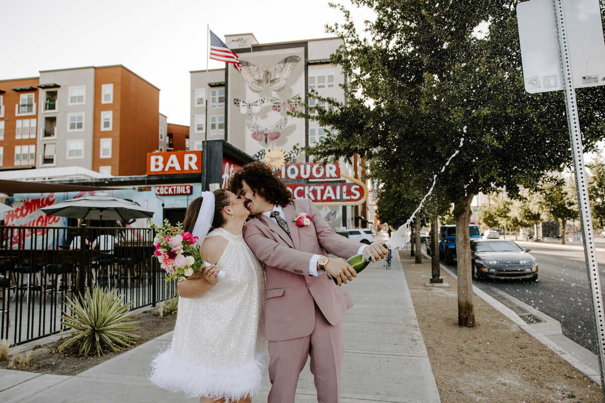 A couple leans in for a kiss while of them pops champagne.