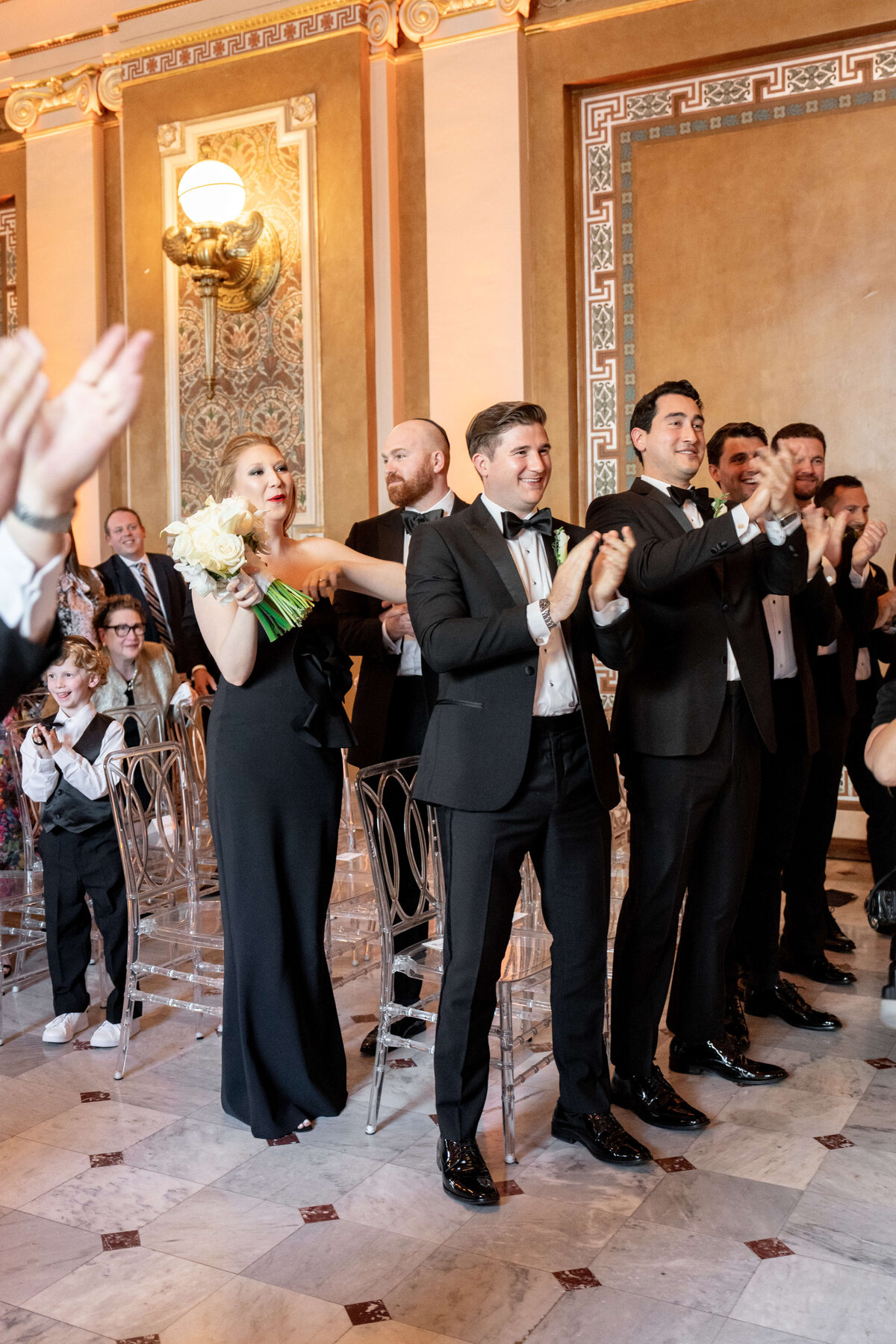 A group of well-dressed people in formal attire clap and smile in an ornate room with decorative walls. A woman in a black dress holds a bouquet while others wear tuxedos. A child seated in front watches the event.