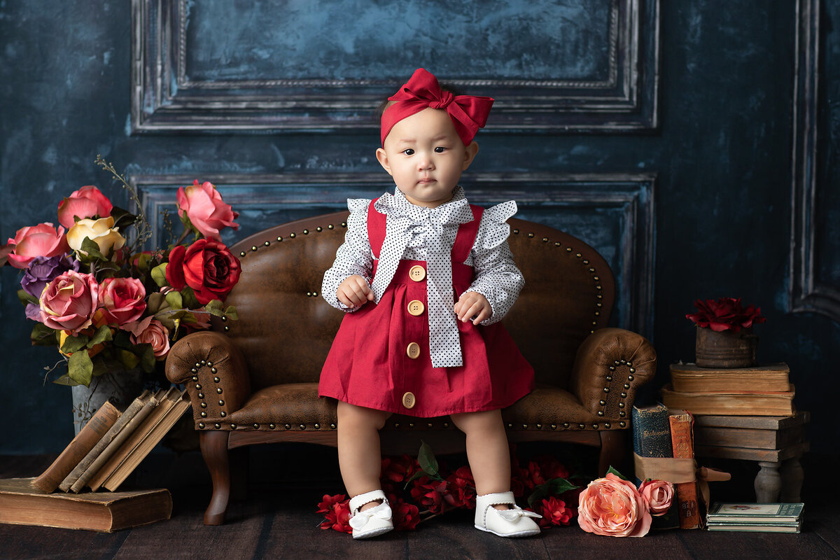 A baby girl dressed in a red jumper and polka dot blouse stands confidently on a vintage leather couch surrounded by books and blooms. This charming setup is a perfect example of Dallas milestone photography that blends elegance with personality.