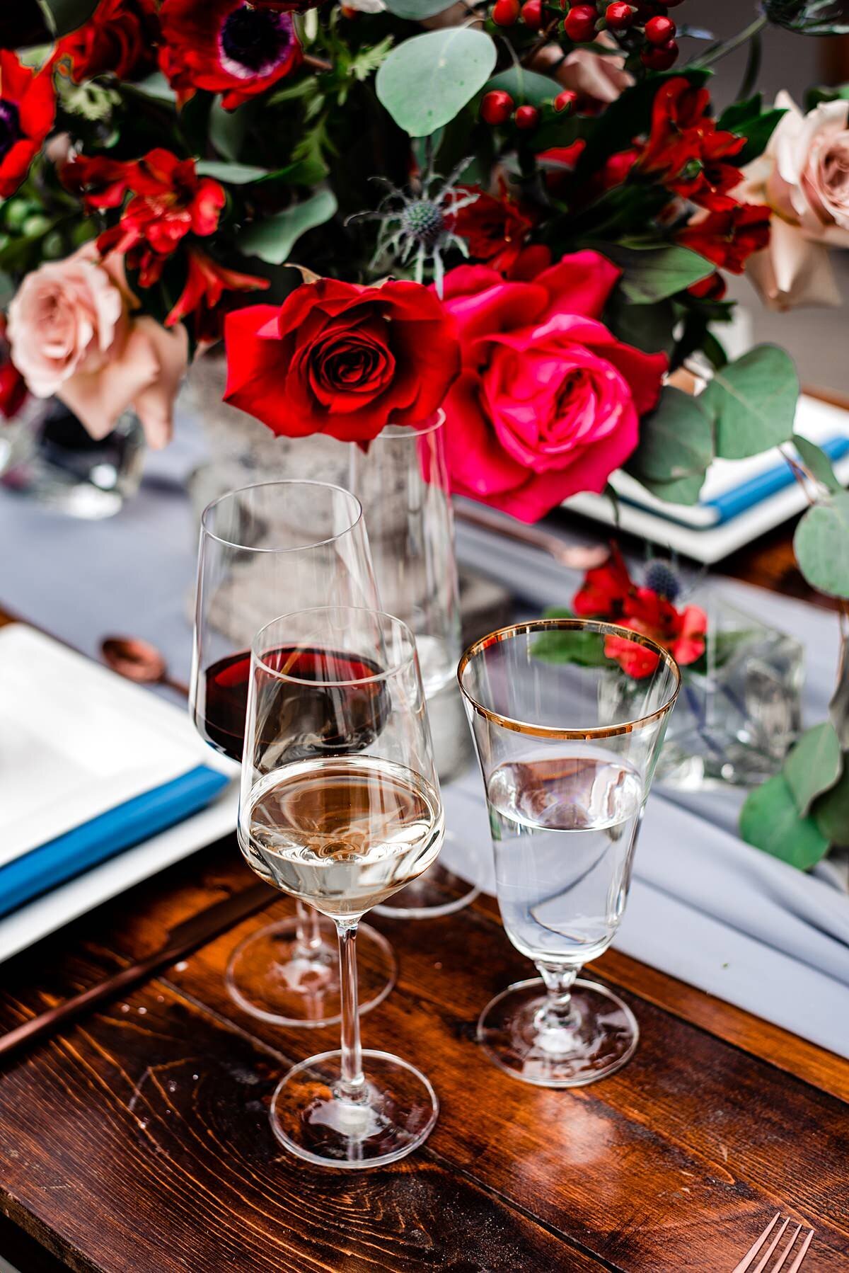 A farm table set with square white plates and a teal linen napkin, a dove gray sheer organza table runner, a large centerpiece of red roses, blush roses, red hypericum berries and blue thistle. There is a large tulip shaped stemmed red wine glass, a smaller stemmed tulip shaped white wine glass and a footed crystal water goblet with a gold rim on the table at Arrington Vineyards.