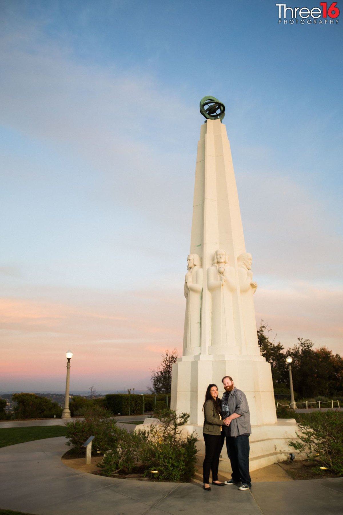 Engaged couple pose together in front of monumental sculpture celebrating astronomers at sunset