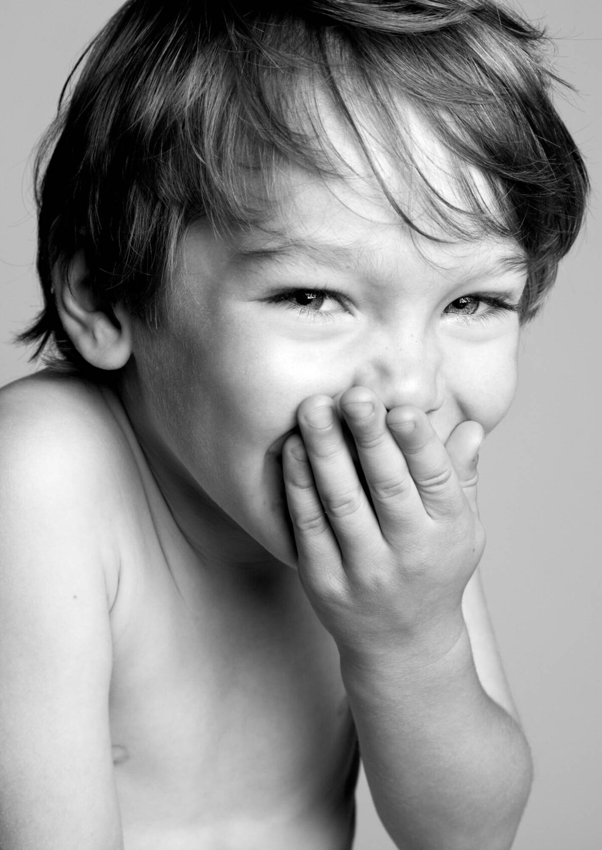 A young boy laughs joyfully while playfully covering his mouth with his hand.