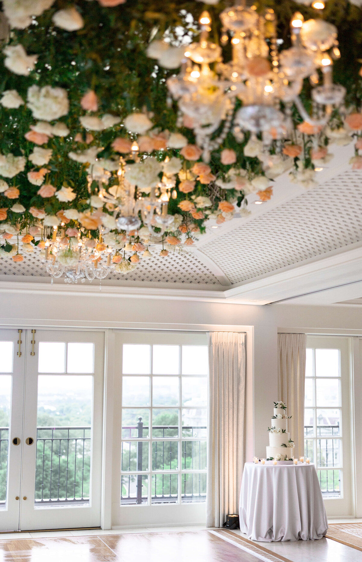 A wedding venue featuring elegant decor with a ceiling adorned with flowers and chandeliers. A three-tiered cake on a table sits in front of large windows with cream-colored curtains, providing a view of lush greenery outside.