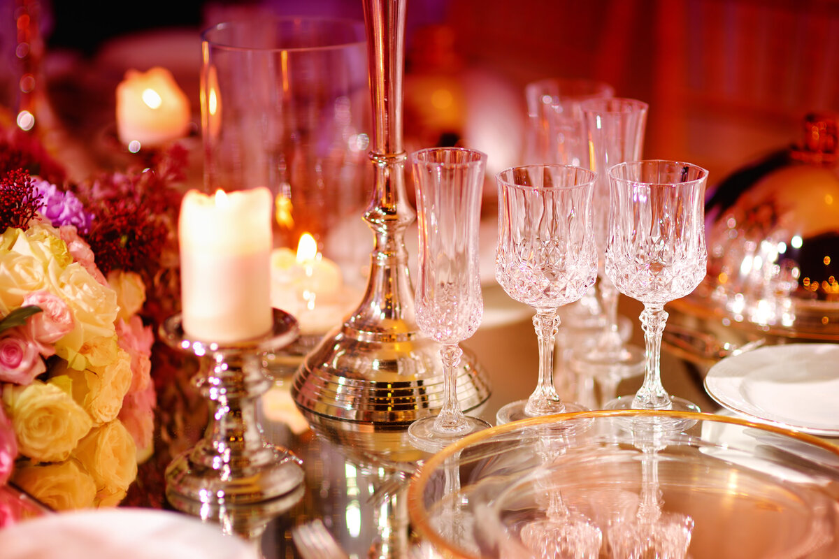 A luxurious wedding tablescape featuring crystal glassware, silver candle holders, and glowing candlelight, creating a warm and romantic ambiance. The rich red glow enhances the elegant centerpieces, adding depth and sophistication to this timeless reception design.