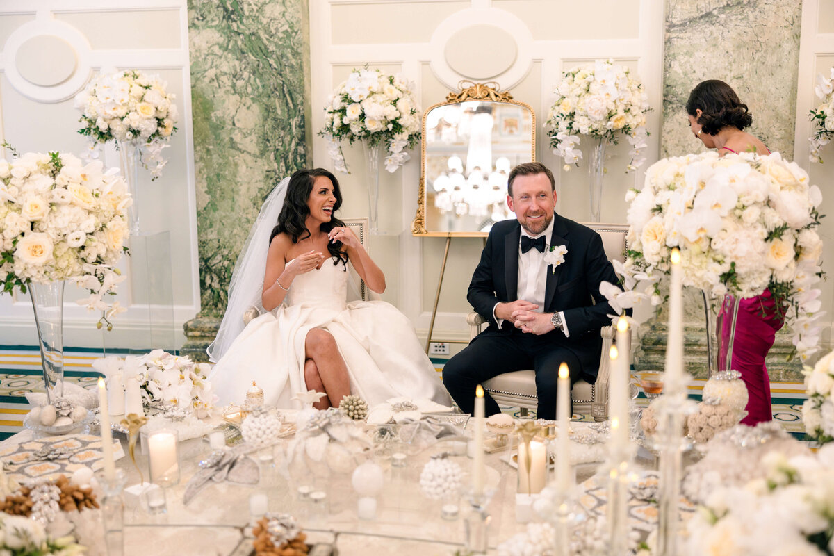 A bride and groom sit in an elegantly decorated room filled with white flowers and lit candles. The bride, in a white gown, and the groom, in a dark suit, are smiling. A mirrored wall and floral arrangements enhance the luxurious setting.