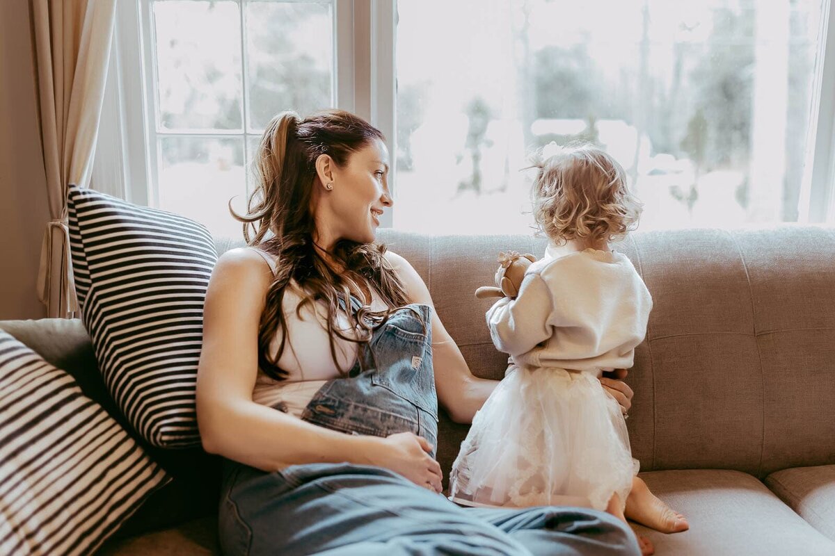 A woman in overalls is sitting on a couch near a window, smiling at a young child standing beside her, who is wearing a white dress and holding a toy.