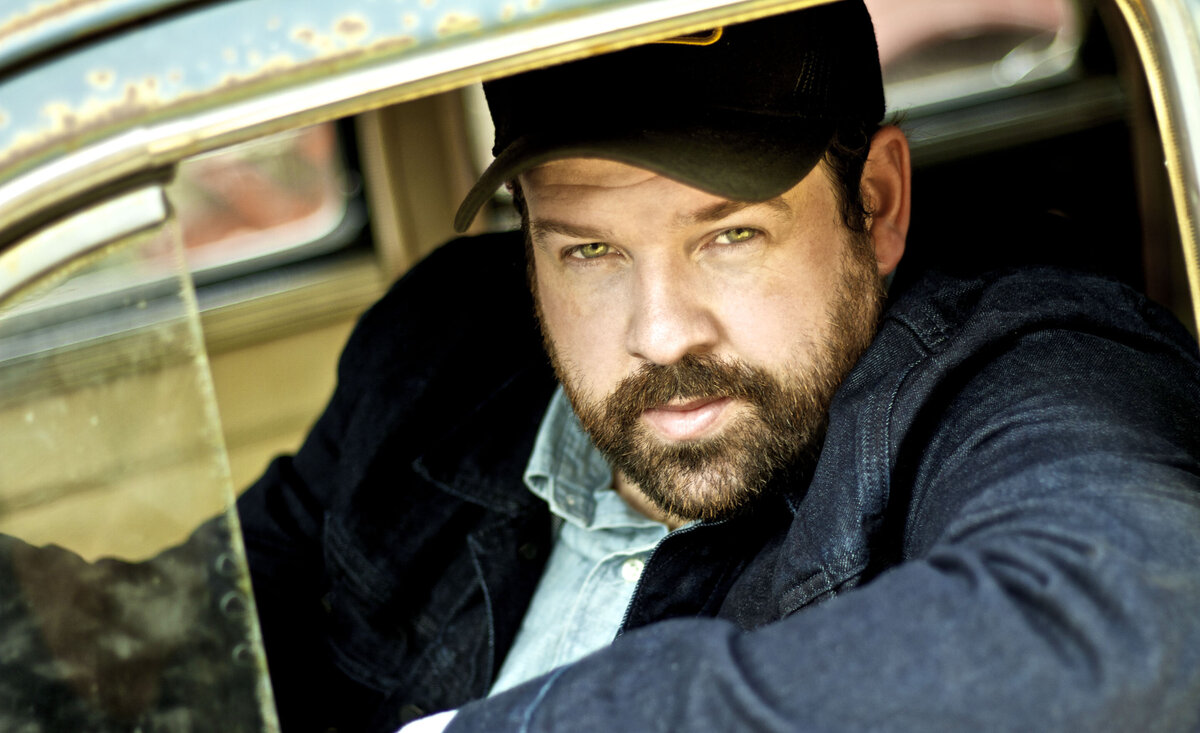 Male musician photo Donovan Woods wearing baseball hat denim coat leaning out old car window