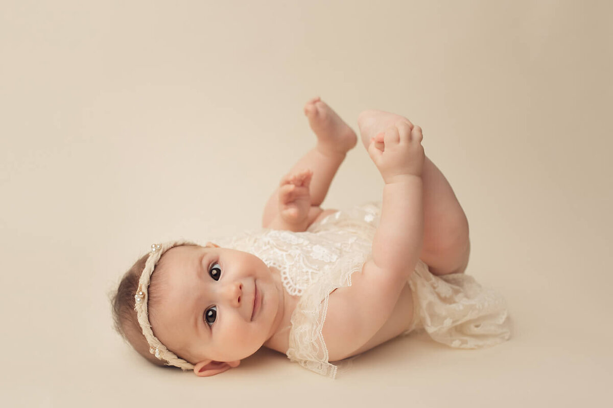 baby girl on her back holding her toes looking at the camera in a white dress
