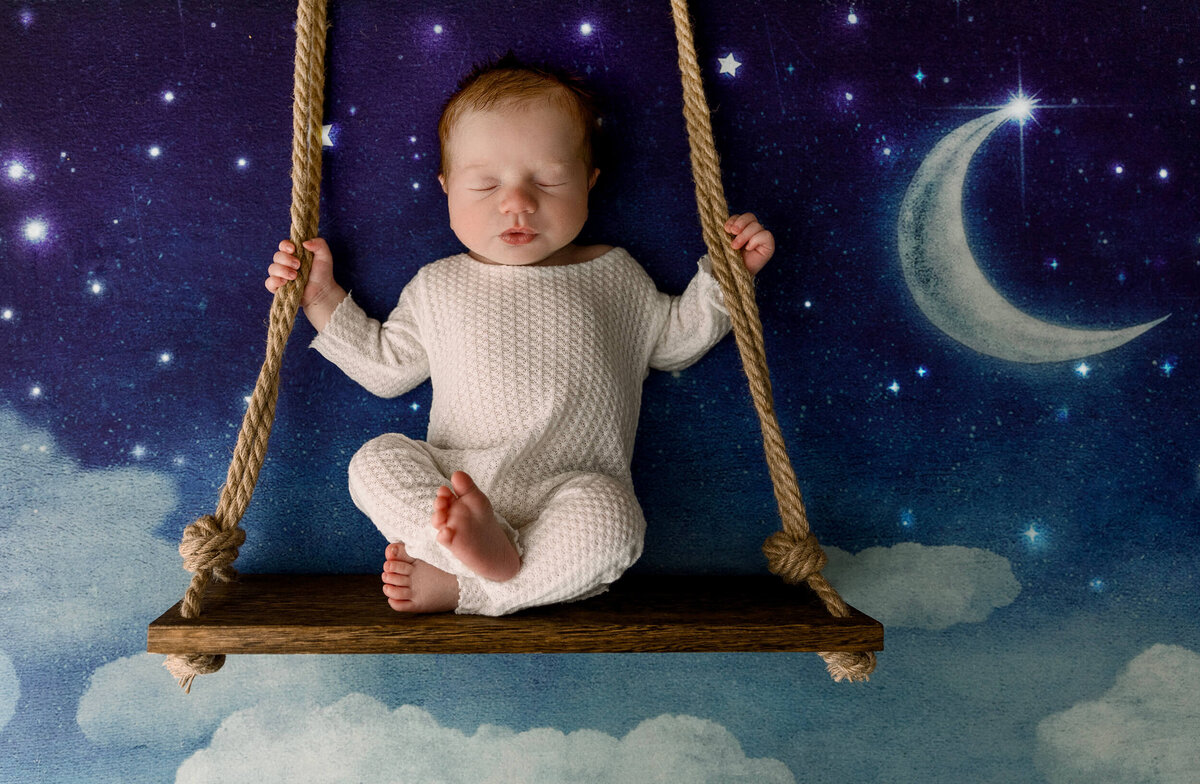Newborn baby boy in a white sleeper on a swing with moon and stars in background