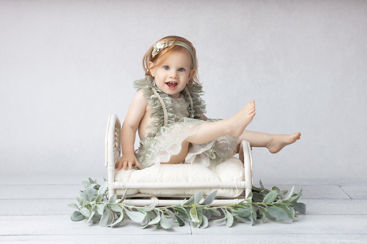 Smiling toddler in a lacy sage green dress playfully lifting her leg while sitting on a mini white bed, surrounded by soft greenery, captured by a DFW milestone photographer.