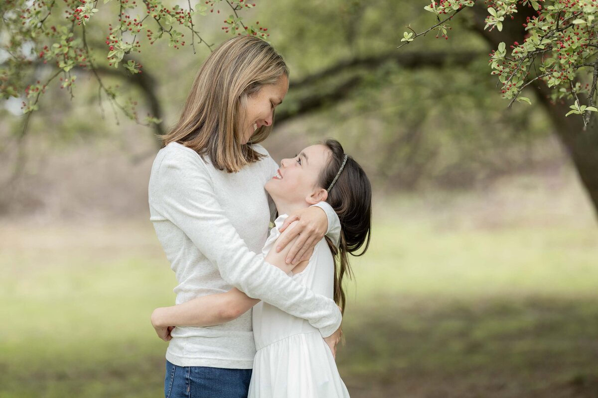 Motherhood photo of a mom and daughter smiling and embracing beneath a flowering tree in Spring 